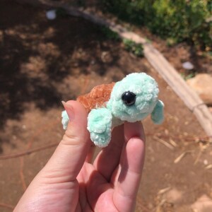 Puede incluir: Una peque&ntilde;a tortuga de peluche hecha a mano con cabeza y extremidades de color verde claro y caparaz&oacute;n marr&oacute;n. La tortuga tiene un ojo negro y est&aacute; sostenida en la mano de una persona. El peluche es un art&iacute;culo lindo y caprichoso.