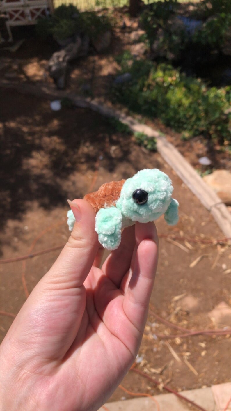 Puede incluir: Una peque&ntilde;a tortuga de peluche hecha a mano con cabeza y extremidades de color verde claro y caparaz&oacute;n marr&oacute;n. La tortuga tiene un ojo negro y est&aacute; sostenida en la mano de una persona. El peluche es un art&iacute;culo lindo y caprichoso.
