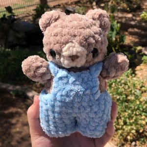 Op de afbeelding: Een handgemaakte, pluche teddybeer met bruine vacht en een lichtblauwe overall. De beer heeft zwarte ogen en een cr&egrave;mekleurige snuit. De beer is ongeveer 15 cm hoog.
