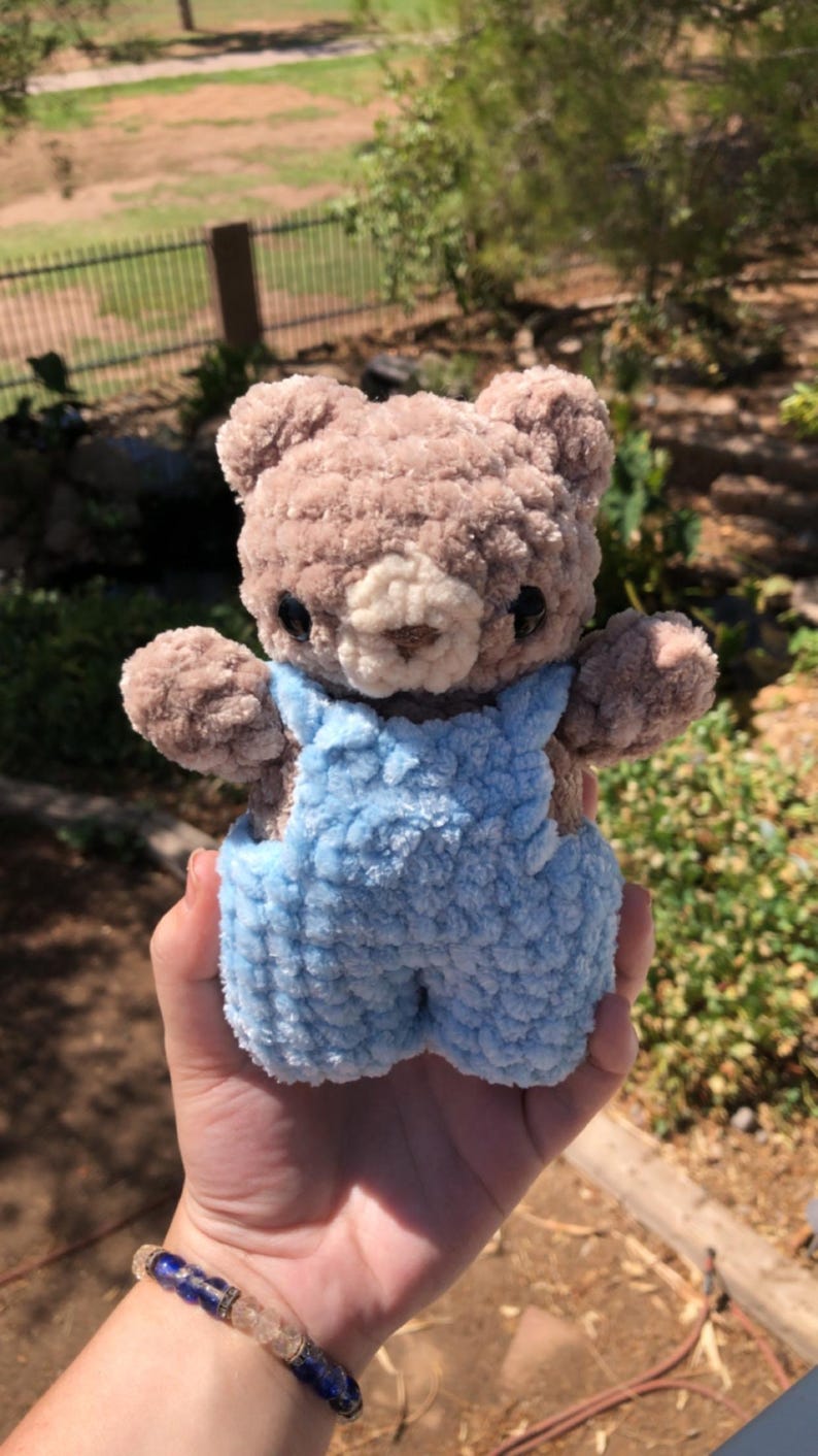 Op de afbeelding: Een handgemaakte, pluche teddybeer met bruine vacht en een lichtblauwe overall. De beer heeft zwarte ogen en een cr&egrave;mekleurige snuit. De beer is ongeveer 15 cm hoog.