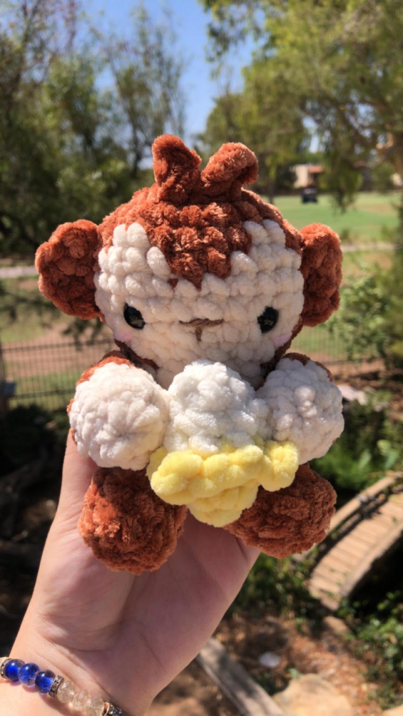 May include: A handmade crochet monkey plushie in brown, white, and yellow. The monkey has large ears, a smiling face, and is holding a yellow and white object. The plushie is soft and cuddly, perfect for a child's toy or nursery decor.