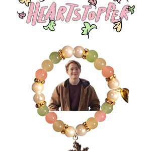 K&ouml;nnte beinhalten: Ein pastellfarbenes Armband in Rosa, Gr&uuml;n und Wei&szlig; mit einem silbernen Ahornblatt-Anh&auml;nger. Das Armband hat ein Foto einer Person in einer braunen Jacke im Kreis der Perlen. Der Text "HEARTSTOPPER" ist in Rosa und Gr&uuml;n &uuml;ber dem Armband geschrieben.
