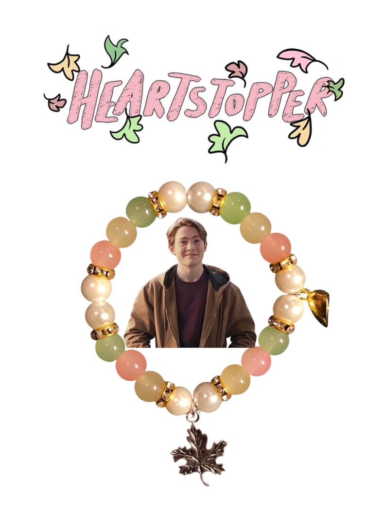 K&ouml;nnte beinhalten: Ein pastellfarbenes Armband in Rosa, Gr&uuml;n und Wei&szlig; mit einem silbernen Ahornblatt-Anh&auml;nger. Das Armband hat ein Foto einer Person in einer braunen Jacke im Kreis der Perlen. Der Text "HEARTSTOPPER" ist in Rosa und Gr&uuml;n &uuml;ber dem Armband geschrieben.
