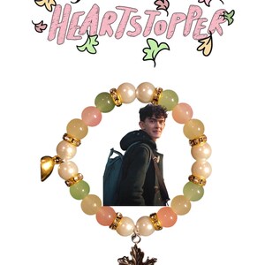K&ouml;nnte beinhalten: Ein Armband mit gr&uuml;nen, rosa und wei&szlig;en Perlen, einem goldenen Anh&auml;nger und einem silbernen Ahornblatt-Anh&auml;nger. Das Armband befindet sich auf einem wei&szlig;en Hintergrund mit dem Text "HEARTSTOPPER" in rosa und gr&uuml;n.