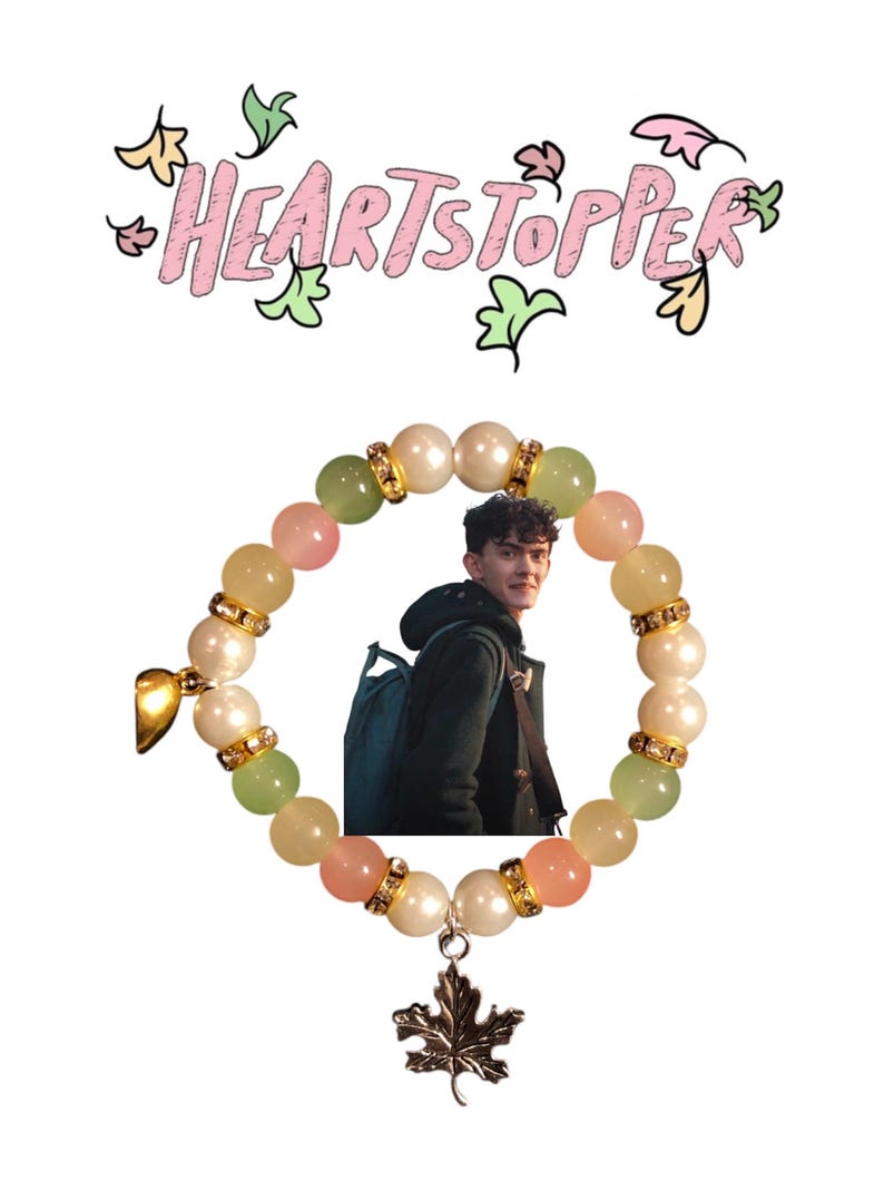 K&ouml;nnte beinhalten: Ein Armband mit gr&uuml;nen, rosa und wei&szlig;en Perlen, einem goldenen Anh&auml;nger und einem silbernen Ahornblatt-Anh&auml;nger. Das Armband befindet sich auf einem wei&szlig;en Hintergrund mit dem Text "HEARTSTOPPER" in rosa und gr&uuml;n.