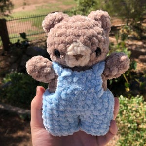 Op de afbeelding: Een handgemaakte gehaakte teddybeer met een lichtbruin lichaam en een lichtblauwe overall. De beer heeft zwarte ogen en een beige snuit. Het pluche speelgoed wordt in een hand gehouden tegen een vage buitenachtergrond.