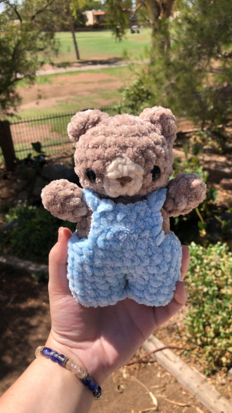Op de afbeelding: Een handgemaakte gehaakte teddybeer met een lichtbruin lichaam en een lichtblauwe overall. De beer heeft zwarte ogen en een beige snuit. Het pluche speelgoed wordt in een hand gehouden tegen een vage buitenachtergrond.