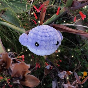 May include: A handmade, plush, lavender whale with white accents and black eyes. The crocheted whale is set against a backdrop of green foliage, red flowers, and brown dried plants. The whale is a whimsical, handcrafted item.