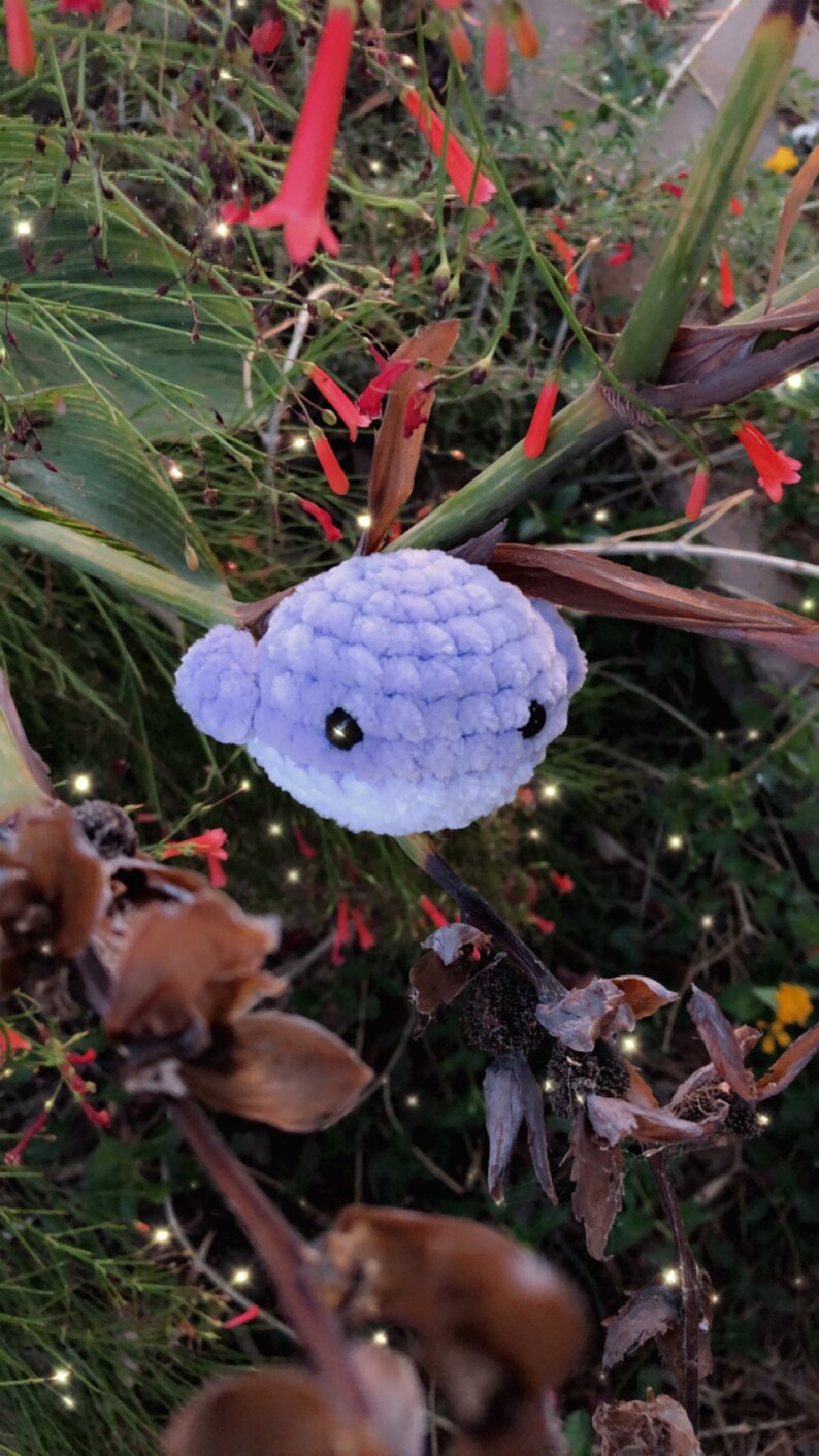 May include: A handmade, plush, lavender whale with white accents and black eyes. The crocheted whale is set against a backdrop of green foliage, red flowers, and brown dried plants. The whale is a whimsical, handcrafted item.