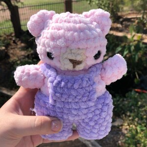 Op de afbeelding: Een handgemaakte gehaakte teddybeer in roze en paars. De beer heeft een lichtroze hoofd, armen en benen, met een donkerpaarse overall. Hij heeft kleine zwarte ogen en een bruine neus. De beer is een zacht, pluche speelgoed.