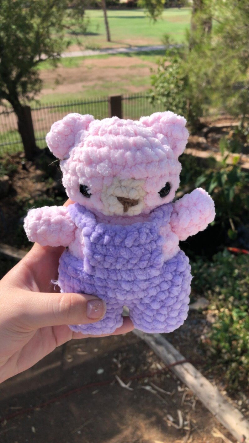 Op de afbeelding: Een handgemaakte gehaakte teddybeer in roze en paars. De beer heeft een lichtroze hoofd, armen en benen, met een donkerpaarse overall. Hij heeft kleine zwarte ogen en een bruine neus. De beer is een zacht, pluche speelgoed.