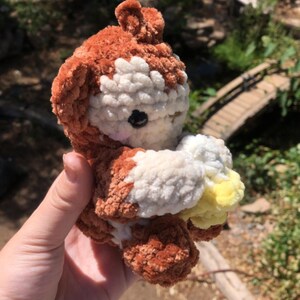 May include: A handmade crochet plush toy in the shape of a bear. The bear is primarily brown with white and yellow accents. It has a black eye and is held in a person's hand.