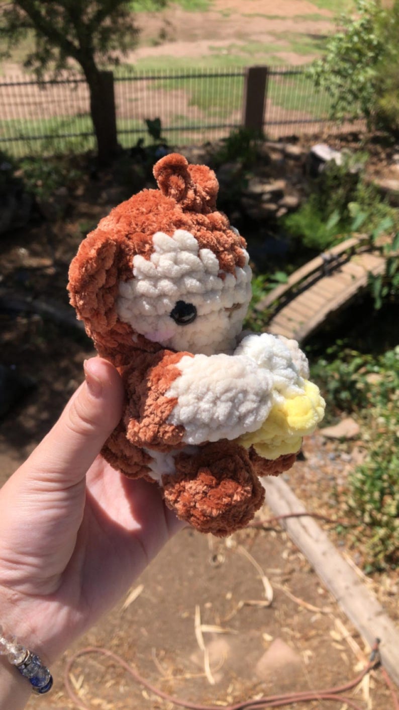 May include: A handmade crochet plush toy in the shape of a bear. The bear is primarily brown with white and yellow accents. It has a black eye and is held in a person's hand.