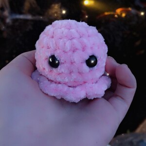 May include: A small, plush pink octopus with large black eyes. The crocheted toy is held in a hand, with a blurred background of a pond and foliage. The octopus has a rounded head and eight small tentacles.