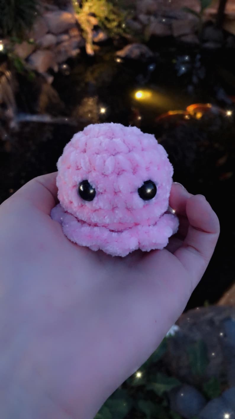 May include: A small, plush pink octopus with large black eyes. The crocheted toy is held in a hand, with a blurred background of a pond and foliage. The octopus has a rounded head and eight small tentacles.