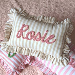 May include: A rectangular throw pillow featuring a beige and white vertical striped pattern with a ruffled border. The name Rosie is embroidered in pink yarn using a textured punch needle technique across the centre of the pillow surface.