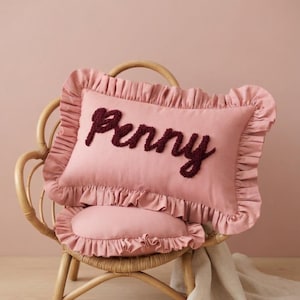 May include: A rectangular dusty pink cushion featuring a ruffled border and the name Penny embroidered in dark maroon yarn across the centre. The cushion is constructed with a soft fabric finish and displays a textured, raised script lettering style.
