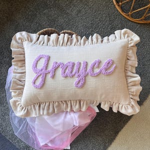 May include: A rectangular beige throw pillow featuring a ruffled border and the name Grayce embroidered in a raised, textured lavender script font across the centre. The pillow is positioned above a piece of white and pink sheer fabric.