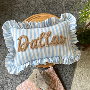 May include: A rectangular throw pillow featuring a light blue and white vertical striped pattern with a ruffled border. The centre of the pillow displays the name Dallas in a raised, textured, tan-coloured yarn script embroidery.