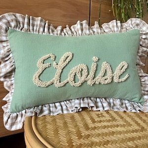 May include: A rectangular sage green throw pillow featuring a ruffled border in a beige and white gingham pattern. The centre of the pillow displays the name Eloise embroidered in a raised, textured cream-coloured yarn script font.