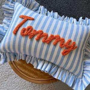 May include: A rectangular throw cushion featuring a light blue and white vertical striped pattern with a ruffled border. The name Tommy is embroidered in the centre using textured, bright orange yarn in a cursive, punch needle style script.
