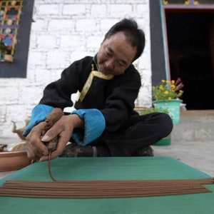 May include: A person is sitting on the ground and making incense sticks. They are using their hands to roll the incense sticks on a green mat. The incense sticks are brown and are arranged in a row on the mat.