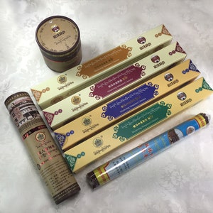 Puede incluir: Una colección de seis inciensos en varios colores y embalajes. El embalaje presenta un diseño dorado y rojo con texto tibetano y las palabras "Incienso del Monasterio de Mindeling".