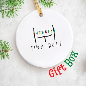 May include: White ceramic Christmas ornament with a black line drawing of a bed with string lights hanging above it. The text "TINY BUTT" is written below the bed.