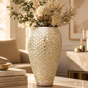 May include: A decorative vase with a mosaic pattern, filled with white and green floral arrangements. The vase has a pearlescent finish and sits on a marble-effect surface. The arrangement includes white hydrangeas and green eucalyptus.