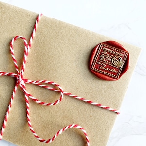 Puede incluir: Un sobre de papel marr&oacute;n sellado con un sello de cera rojo y atado con hilo rojo y blanco. El sello de cera tiene letras doradas y un dise&ntilde;o circular. El hilo est&aacute; atado en un lazo decorativo.