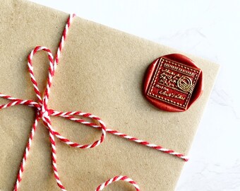 Op de afbeelding: Een bruine papieren envelop verzegeld met een rode wasstempel en vastgebonden met rood en wit touw. De wasstempel heeft gouden letters en een cirkelvormig ontwerp. Het touw is in een decoratieve strik geknoopt.