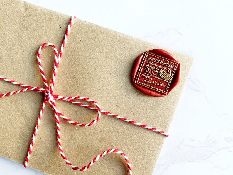 Puede incluir: Un sobre de papel marr&oacute;n sellado con un sello de cera rojo y atado con hilo rojo y blanco. El sello de cera tiene letras doradas y un dise&ntilde;o circular. El hilo est&aacute; atado en un lazo decorativo.