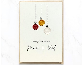 Op de afbeelding: Een crèmekleurige wenskaart met drie hangende waszegelornamenten in rood, goud en wit, opgehangen aan zwarte stippellijnen. De tekst merry christmas en Mum & Dad staat in zwarte inkt op de kaart, die een bruine enveloprand heeft.