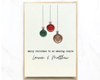 Op de afbeelding: Een crèmekleurige wenskaart met drie hangende waszegelornamenten in groen, rood en koper. De groene zegel heeft een krans, de rode een hart en de koperen een hert. De tekst luidt: merry christmas to an amazing couple Lauren & Matthew.