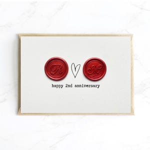 May include: A white card with a brown envelope features two red wax seals with initials and a hand-drawn heart. The text "happy 2nd anniversary" is printed below the seals. The card is on a white marble surface.