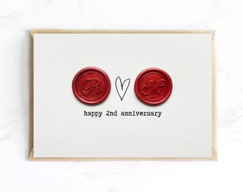 Op de afbeelding: Een witte kaart met een bruine envelop toont twee rode zegels met initialen en een handgetekend hart. De tekst "happy 2nd anniversary" staat eronder gedrukt. De kaart ligt op een wit marmeren oppervlak.