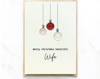 Op de afbeelding: Een crèmekleurige wenskaart met drie hangende ornamenten van waszegels in rood, lichtroze en wit. Het rode zegel heeft een hartje, terwijl de andere florale patronen hebben. De tekst op de kaart luidt merry christmas beautiful Wife.