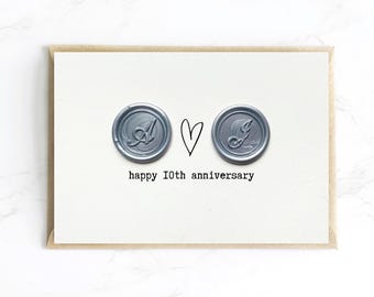 Op de afbeelding: Een wenskaart met een kraftpapier rand. De kaart heeft twee zilveren zegels met de letters "A" en "J", een hart en de tekst "happy 10th anniversary" in een eenvoudig lettertype.