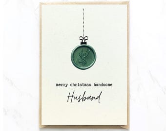 Op de afbeelding: Een crèmekleurige wenskaart met een groen waszegel in de vorm van een kerstbal met een hertenkopontwerp. Een zwarte stippellijn verbindt het zegel met de bovenkant. De tekst luidt merry christmas handsome Husband.