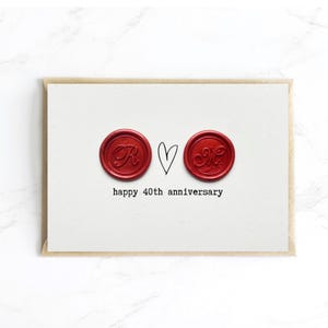 May include: A white card with a brown border features two red wax seals, a heart, and the text "happy 40th anniversary." The seals have the letters "R" and "M" in elegant script. A simple, elegant design.