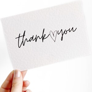 May include: A white card with the words "thank you" in black script, with a heart replacing the "o" in "you". The card is held by a hand against a white background. The card has a textured surface.