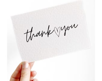 Op de afbeelding: Een witte kaart met de woorden "thank you" in zwart schrift, met een hartje dat de "o" in "you" vervangt. De kaart wordt door een hand tegen een witte achtergrond gehouden. De kaart heeft een getextureerd oppervlak.