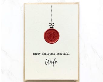 Op de afbeelding: Een kerstkaart met een rood hartvormig ornamentontwerp. Het ornament hangt aan een stippellijn en is bekroond met een strik. De kaart leest "merry christmas beautiful Wife" in zwarte tekst.