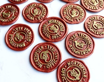 Op de afbeelding: Een verzameling van 12 ronde, rode waszegels met een gouden ontwerp. Elk zegel heeft de woorden "Noordpool" en "Officiële Zegel" in een cirkelvormig ontwerp. De zegels zijn willekeurig gerangschikt op een wit oppervlak.
