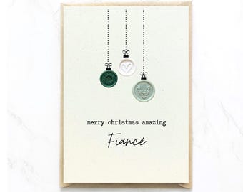 Op de afbeelding: Een kerstkaart met een lichtbeige achtergrond. De kaart heeft drie hangende ornamenten in groen, wit en lichtgroen. De tekst luidt "merry christmas amazing Fiance".
