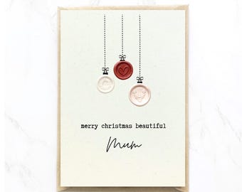 Op de afbeelding: Een crèmekleurige wenskaart met drie hangende waszegelornamenten in rood, wit en lichtroze. De kaart bevat de tekst "merry christmas beautiful" in een typemachinelettertype en het woord "mum" in een zwart handschriftlettertype eronder.
