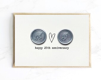 Op de afbeelding: Een wenskaart met een kraft papieren envelop. De kaart toont twee zilveren zegels met de letters "A" en "J", een hart en de tekst "happy 25th anniversary" in een eenvoudig lettertype.