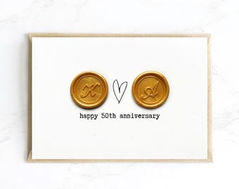Op de afbeelding: Een witte kaart met een kraft papieren envelop heeft twee gouden zegels met de letters "K" en "A", een handgetekend hart en de tekst "happy 50th anniversary" in het zwart. De kaart ligt op een wit marmeren oppervlak.