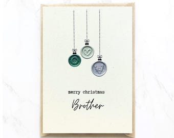 Op de afbeelding: Een kerstkaart met een crèmekleurige achtergrond en een bruine rand. De kaart heeft drie waszegelornamenten in groen, lichtblauw en grijs, hangend aan stippellijnen. De tekst luidt "merry christmas Brother."