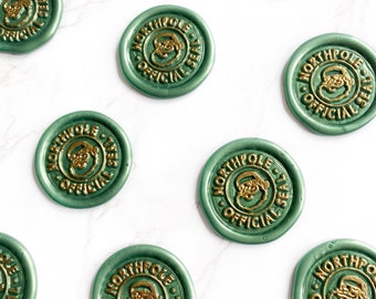 Op de afbeelding: Groene zegels met gouden letters en een ontwerp van de kerstman. Elke zegel leest "NORTH POLE OFFICIAL SEAL" in een cirkelvormig patroon. De zegels zijn verspreid over een wit oppervlak.